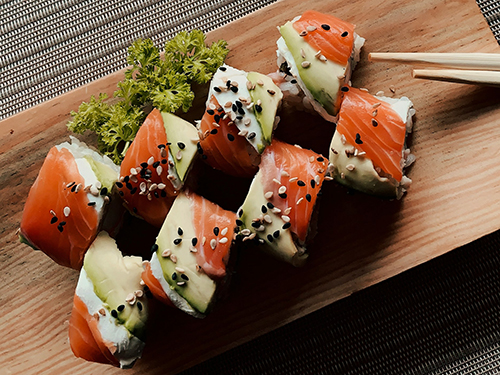 A top-down shot of a wooden board with sushi rolls, garnished with sesame seeds and fresh greens. A small bowl of soy sauce and chopsticks sit beside it.