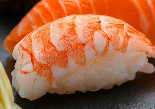 A close-up of beautifully arranged sushi on a dark slate plate. The focus is on the front piece, with the background slightly blurred to highlight the textures and colors.