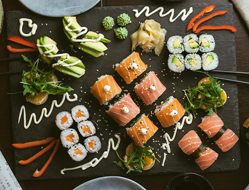 A black serving board covered with sushi rolls, drizzled sauces, fresh greens, and sliced peppers sits on a dark wooden table. Surrounding it are chopsticks, a soy sauce bottle, a bowl of edamame, and a folded newspaper.