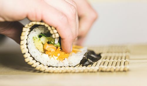 A hand firmly presses a bamboo mat, rolling sushi on a wooden surface. The close-up captures the motion and texture, with the background softly blurred.