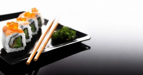 A black plate with a reflective surface holds a row of sushi rolls, with a pair of wooden chopsticks resting beside them. The bright, blurred background contrasts with the dark plate.